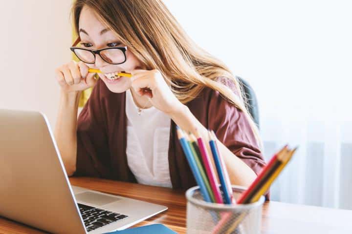 woman biting pencil while sitting on chair in front of computer during daytime learning